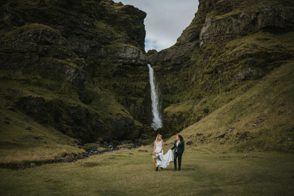 Couple walking and holding hands while smiling at each other with a big waterfall in the background in Iceland.