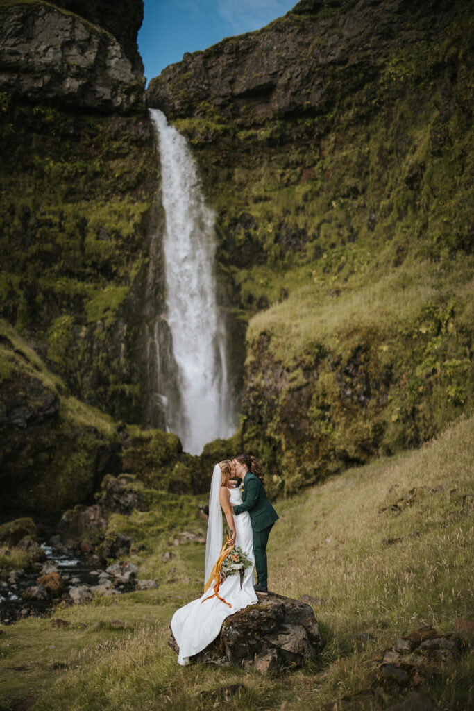 Two brides giving each other a kiss with a beautiful waterfall behind them while they're eloping in Iceland.