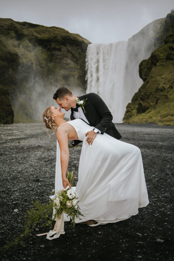 Groom dipping his bride and giving her a big kiss in front of a huge waterfall in Iceland on their elopement wedding day.